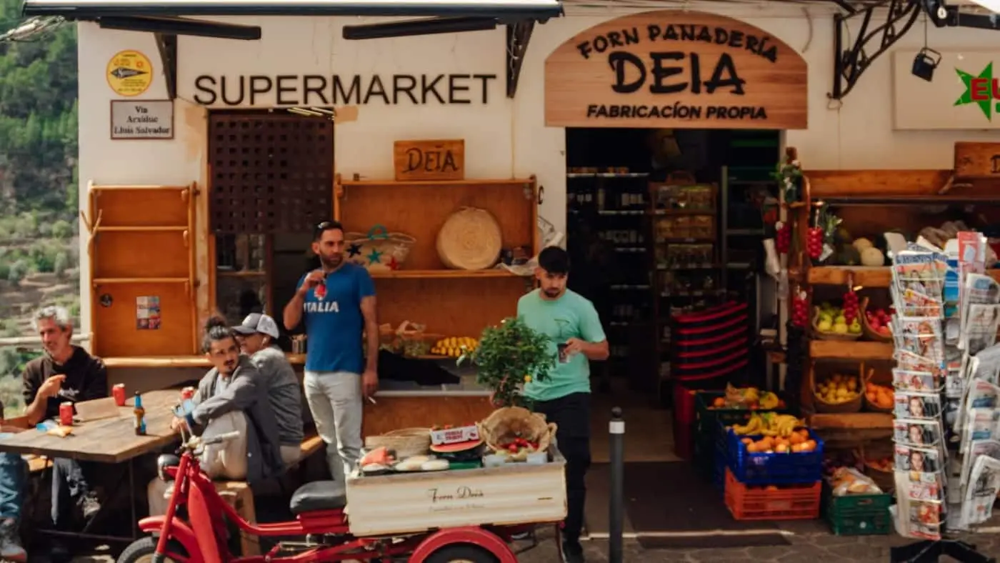Clientes en la puerta de una tienda de barrio con fruta y prensa en la calle